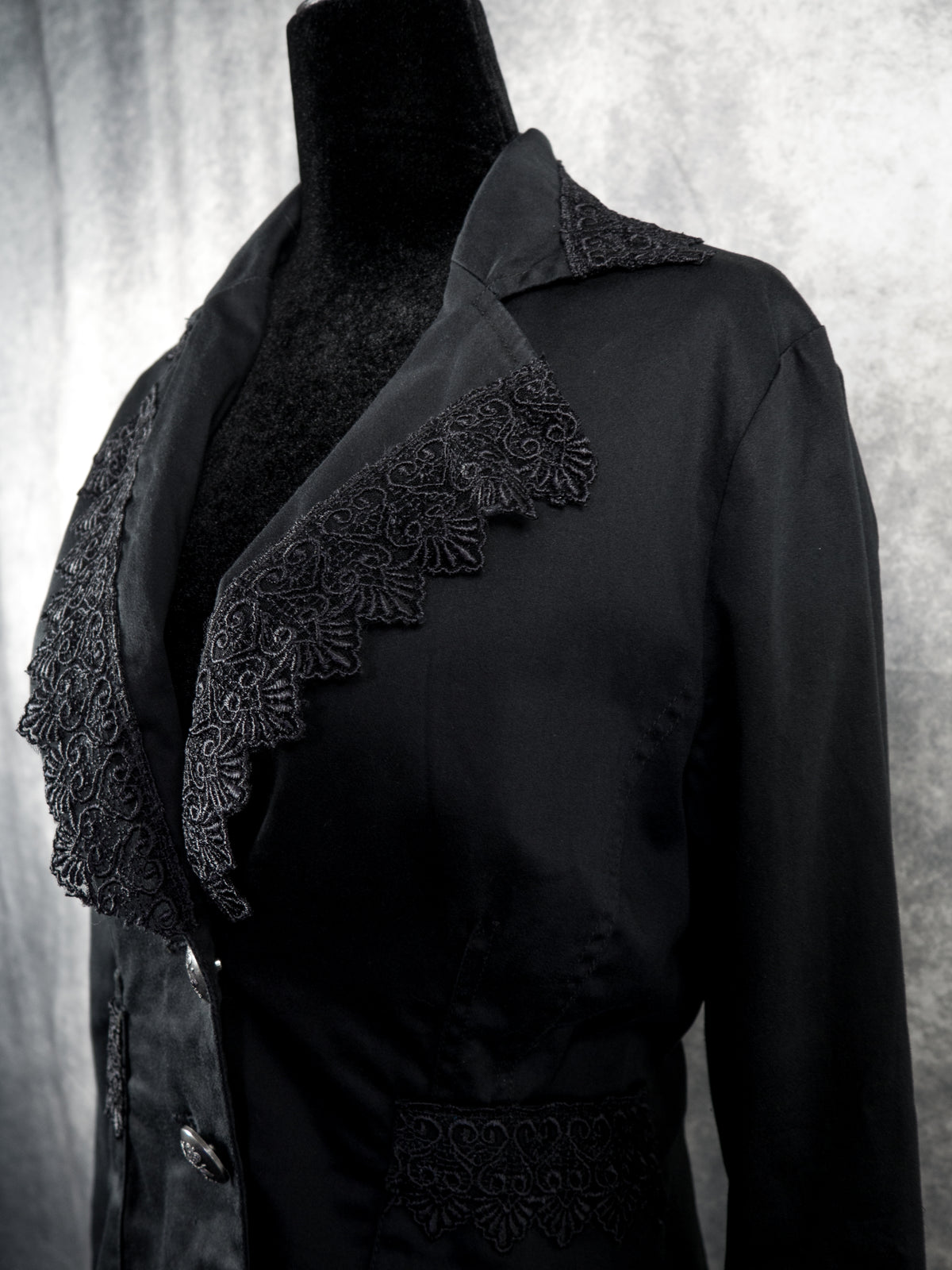 A black long-sleeve blazer displayed on a black mannequin torso, featuring decorative black lace trim along the collar and front opening, against a textured gray studio background.