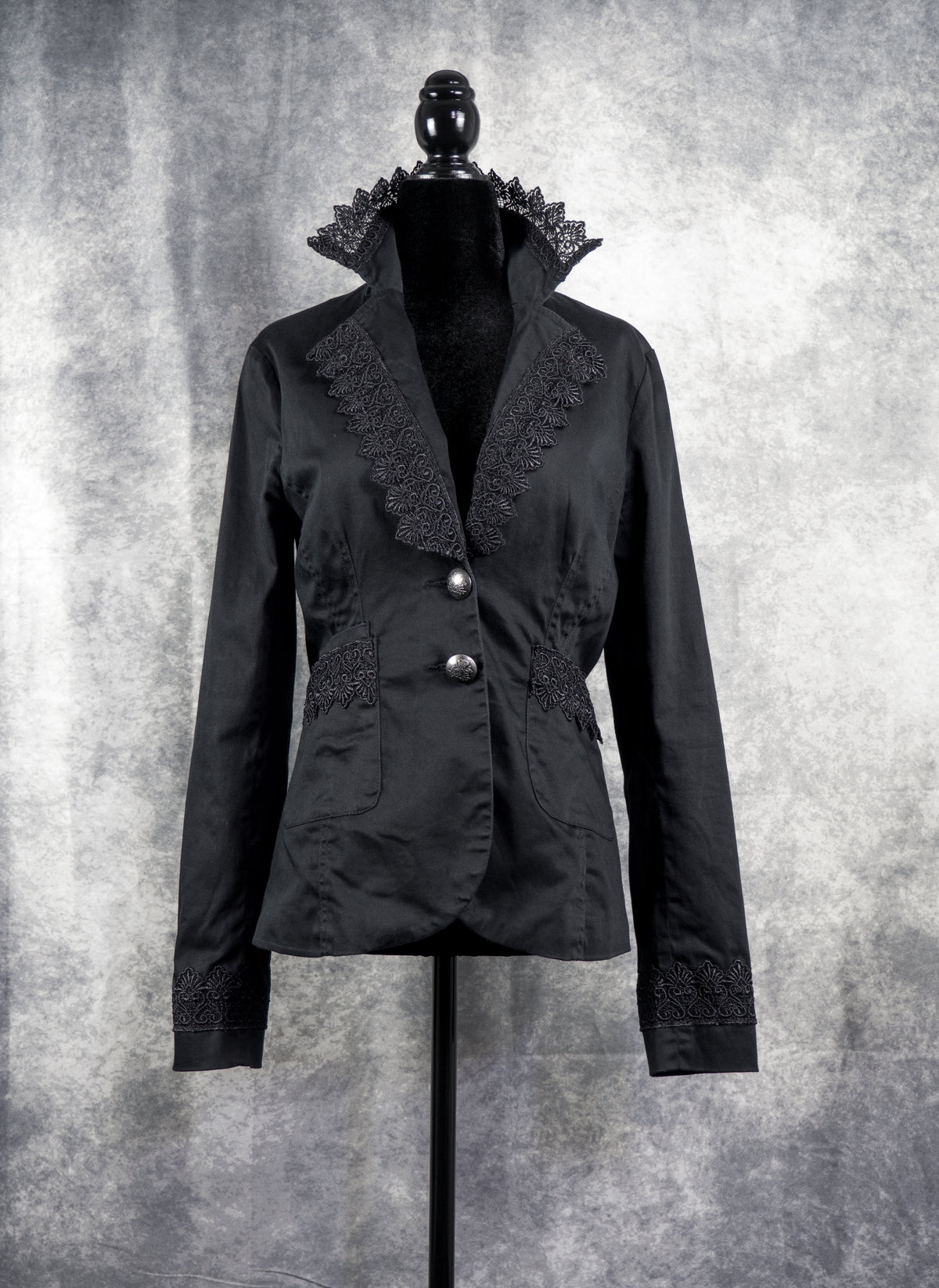  A tailored black alternative blazer displayed on a black female mannequin against a textured grey studio background. The jacket features notched lapels adorned with intricate black floral lace trim along the edges. It secures with two ornate silver-tone statement buttons at the waist, complemented by matching lace details across the side pockets and cuffs to create a gothic, structured silhouette.