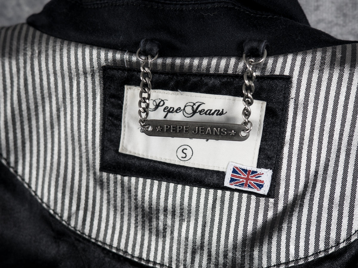 A close-up view of the inner neck area of the black blazer, showing a white fabric label sewn onto a signature Pepe Jeans silver and black pinstriped lining panel. The label features the "Pepe Jeans" logo script, a small silver-tone metal chain loop above a plaque engraved with "★ PEPE JEANS ★", a circle enclosing the letter "S" indicating size small, and a small British Union Jack flag tag stitched in the lower right corner.