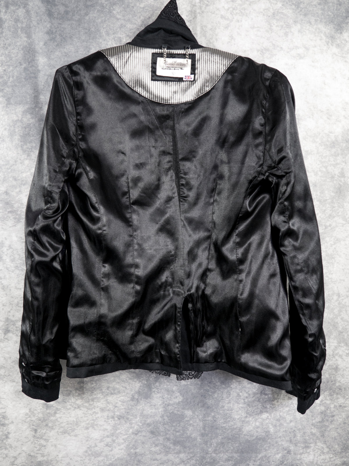 A full view of the inside of the black blazer, showing the smooth black satin interior lining. The top neck area features a contrasting silver and black pinstriped panel with the sewn-in brand label and chain detail. The tailored panels, vertical seams, and structured shoulder construction of the jacket's interior are visible against the grey studio background.
