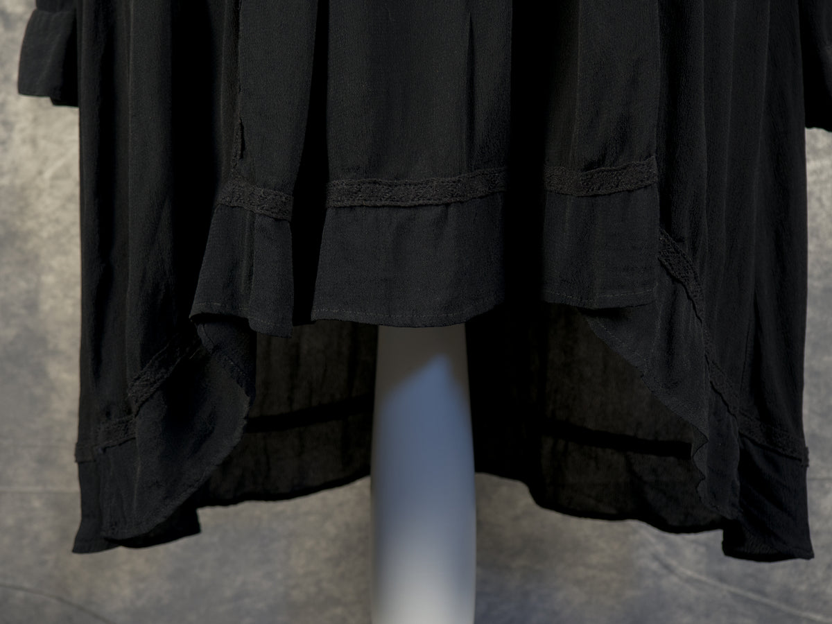 A front close-up view of the high-low hemline on a black dress displayed on a white mannequin stand. The lightweight fabric cascades beautifully, being visibly shorter in the front and longer in the back, accented by a subtle horizontal embroidered lace trim just above the edge.