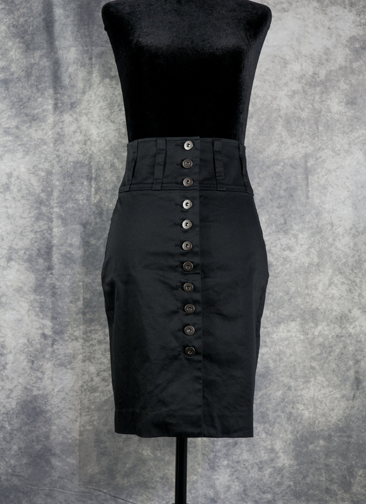 A black high-waisted pencil skirt with a prominent vertical row of dark buttons down the front, displayed on a black dress form against a textured gray background.