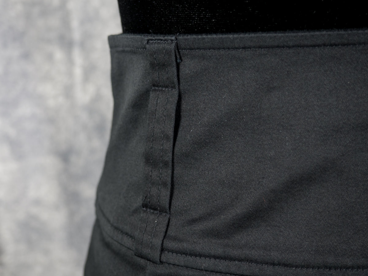 Close-up of a dark charcoal gray garment, showing a vertical seam and a stitched tab detail on the side, with a black waistband at the top against a textured gray background.