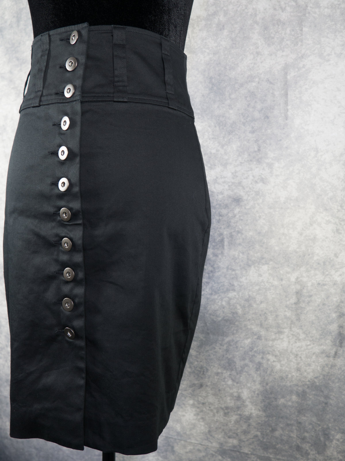 A black high-waisted pencil skirt with a prominent vertical row of silver buttons down the front, displayed on a mannequin torso against a textured gray background.
