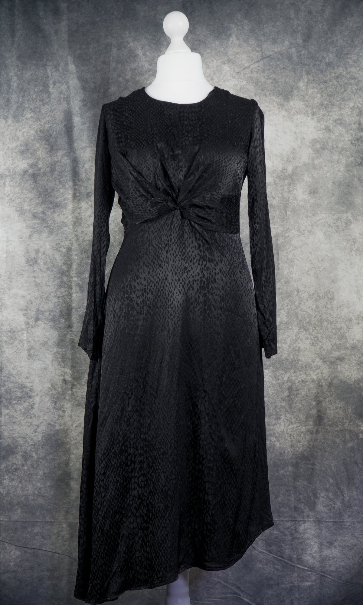A black long-sleeve midi dress with a textured pattern and a twisted knot detail at the waist, displayed on a white mannequin against a mottled gray background.