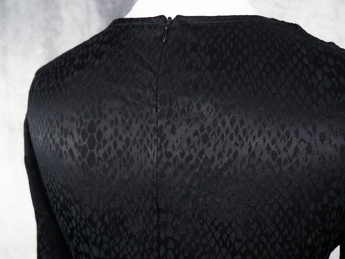 Close-up back view of a black long-sleeve top with a subtle snakeskin pattern and a central zipper, displayed on a mannequin against a light gray textured background.