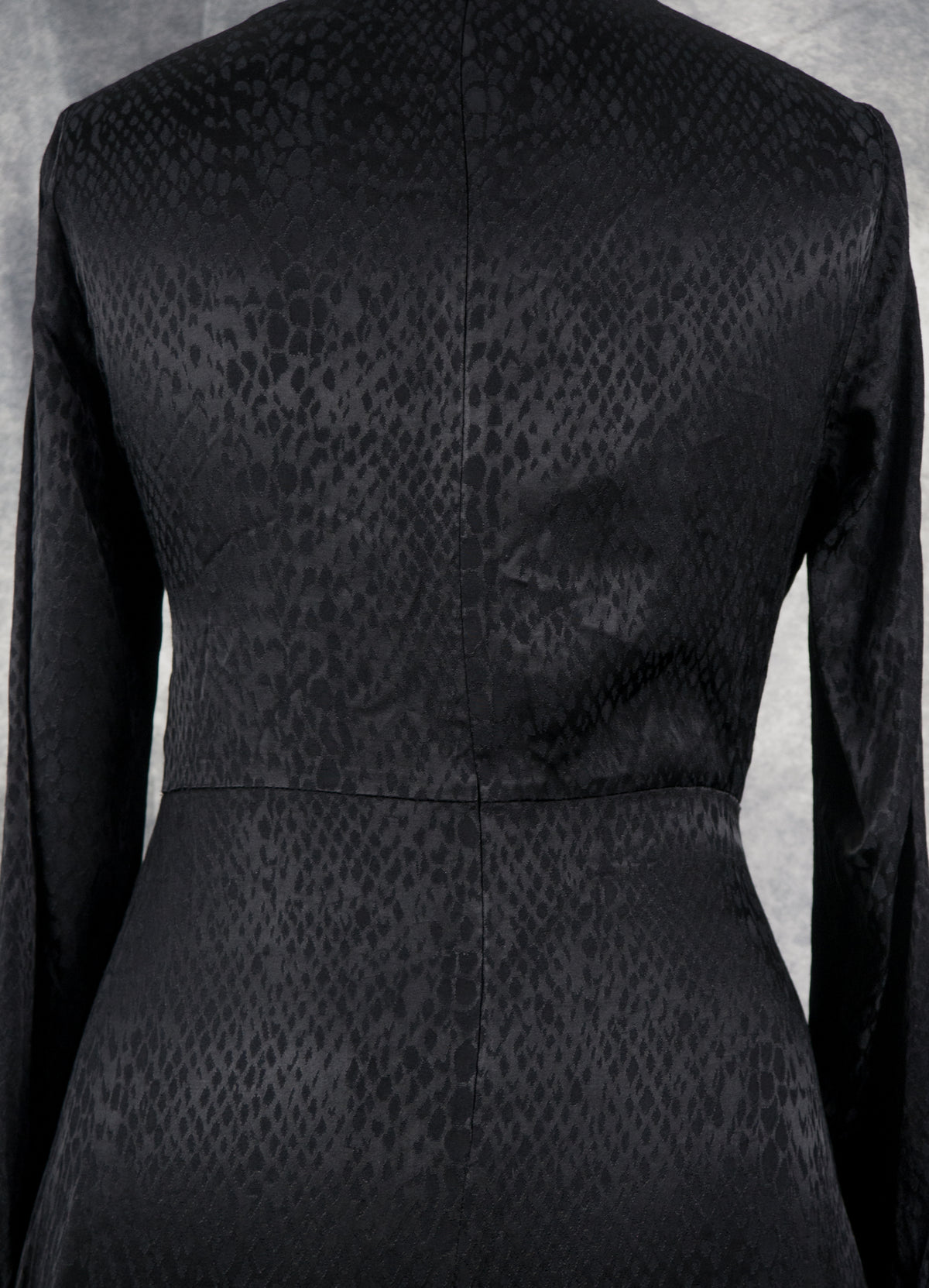 Close-up back view of a black long-sleeve dress with a subtle snakeskin or reptile-embossed pattern, shown against a neutral gray background.