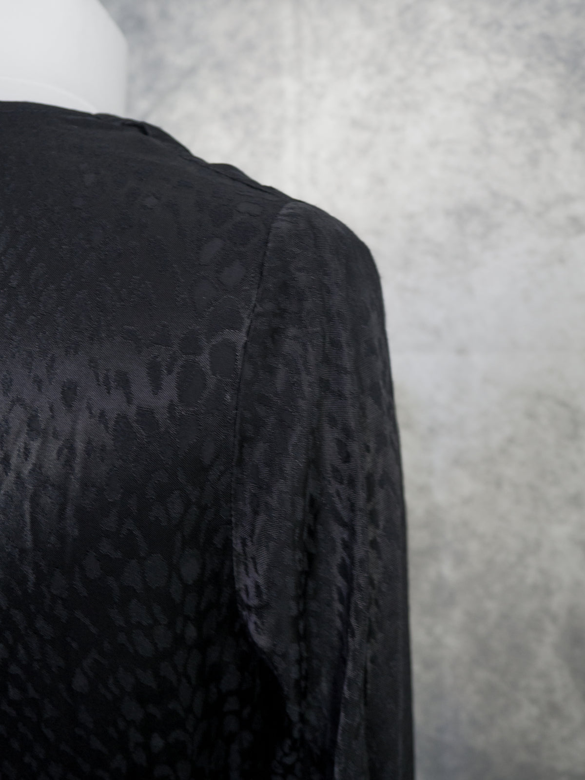 Close-up of a black textured top or dress with a subtle tonal animal-like pattern, shown on a mannequin against a mottled gray background.