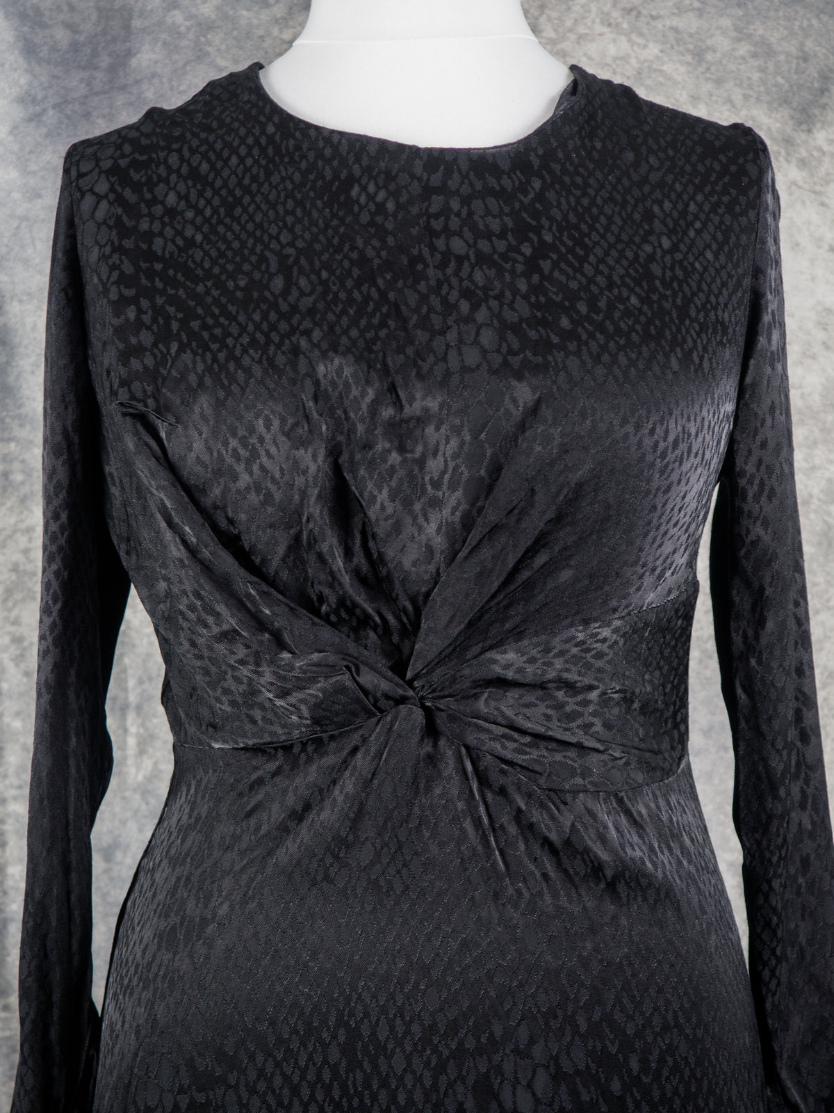 A black long-sleeve dress with a textured snakeskin pattern, featuring a twisted knot detail at the waist, displayed on a white mannequin torso against a gray textured background.