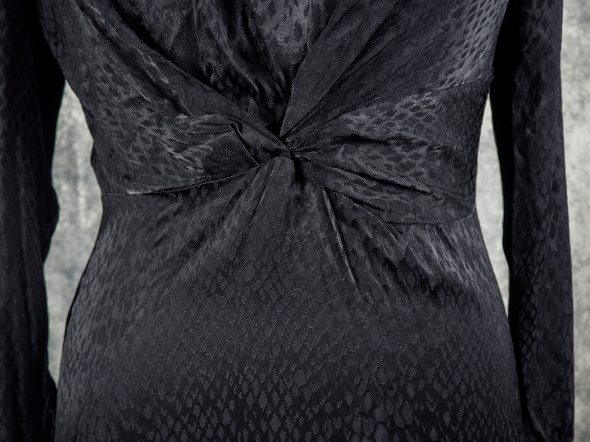 Close-up of a black textured dress with a twisted waist detail and long sleeves, shown on a mannequin against a gray background.