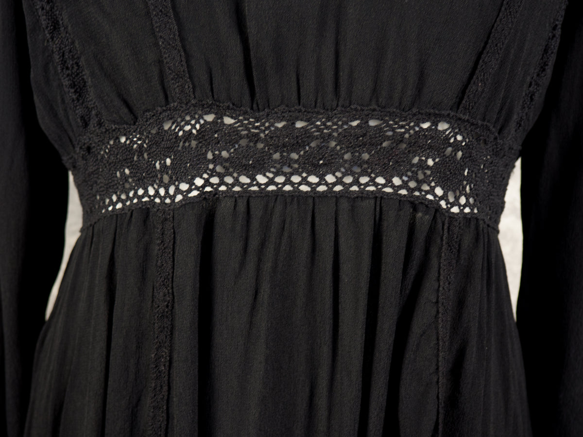 A detailed front close-up of the empire waist on a black dress. The image highlights a horizontal, semi-sheer crocheted lace panel with an openwork geometric design, set between the softly gathered fabric of the bodice and the flowing folds of the skirt.