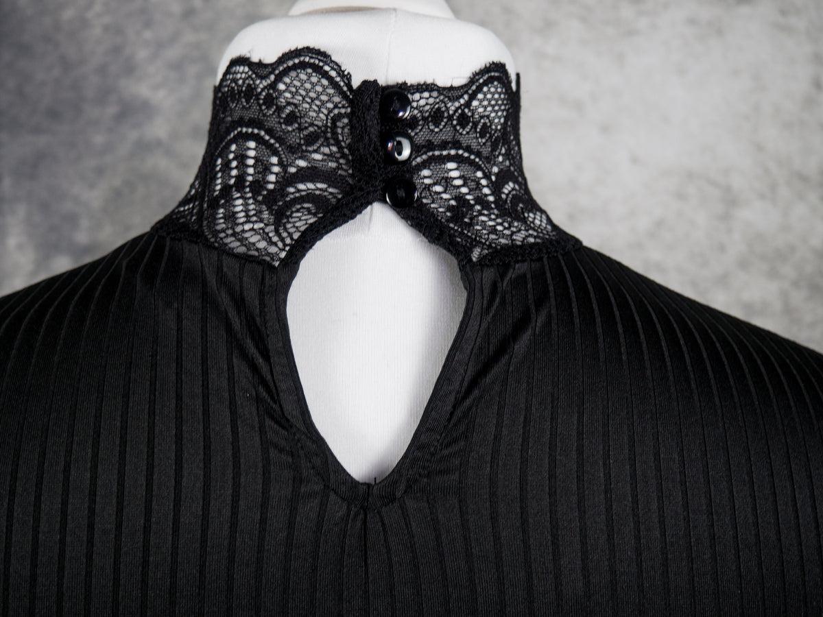 Black pleated top with a high lace-up collar featuring intricate black lace panels and decorative buttons, displayed on a white mannequin torso against a textured gray background.