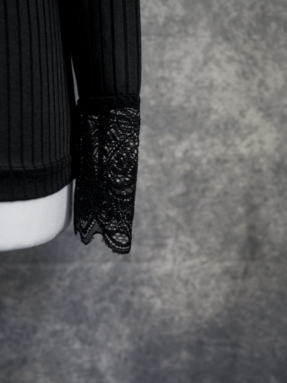 Close-up of a black ribbed knit sleeve with a sheer black lace cuff, displayed on a white mannequin arm against a textured gray background.