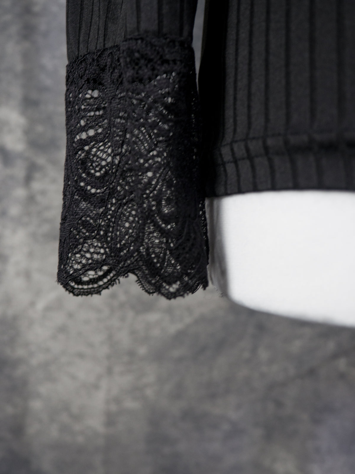 Close-up of a black long-sleeve top featuring a textured ribbed body and a sheer floral lace cuff, displayed on a white mannequin arm against a gray background.