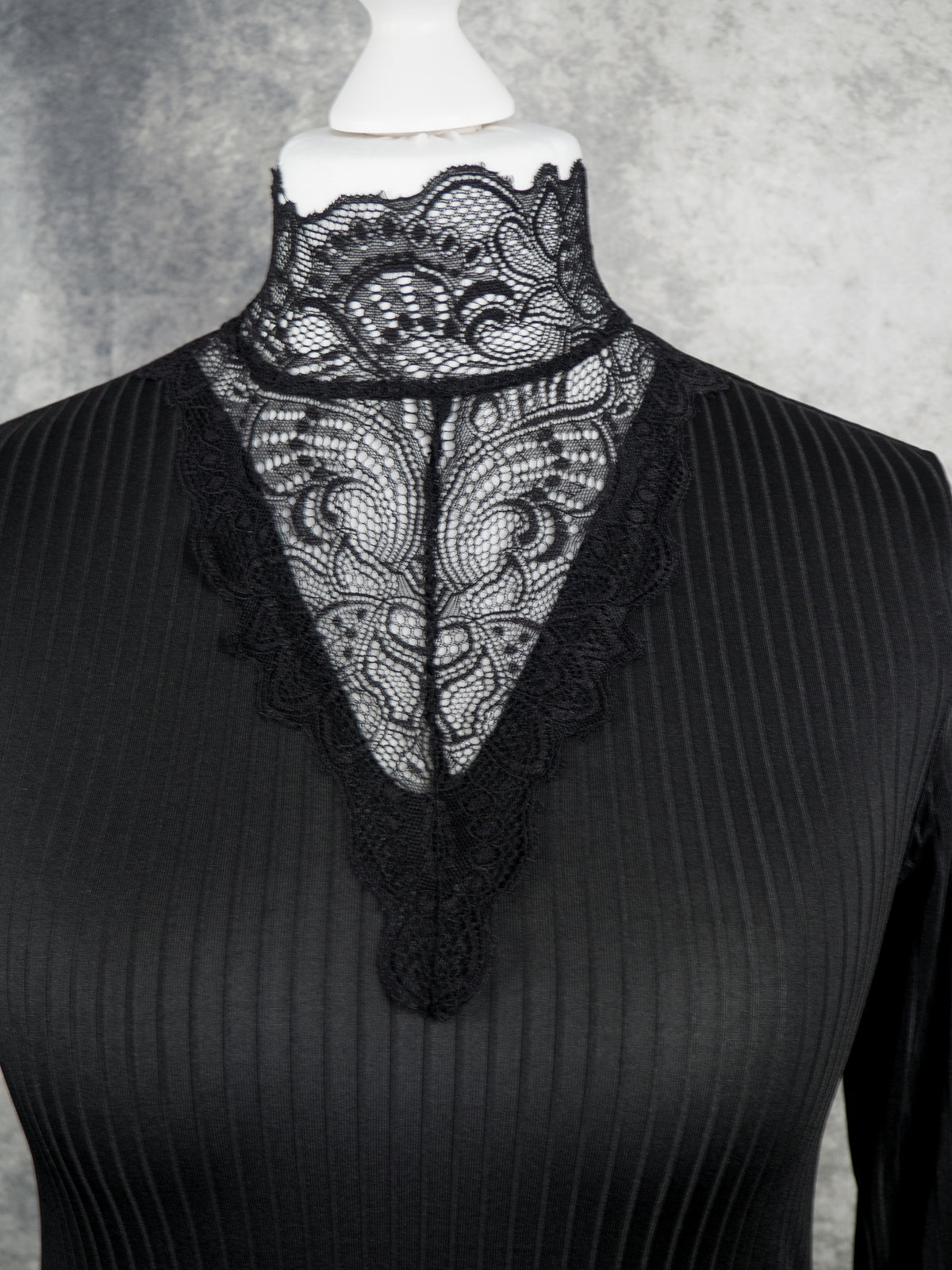 A black ribbed long-sleeve top with a sheer black lace V-neckline panel, displayed on a white mannequin torso against a textured gray background.
