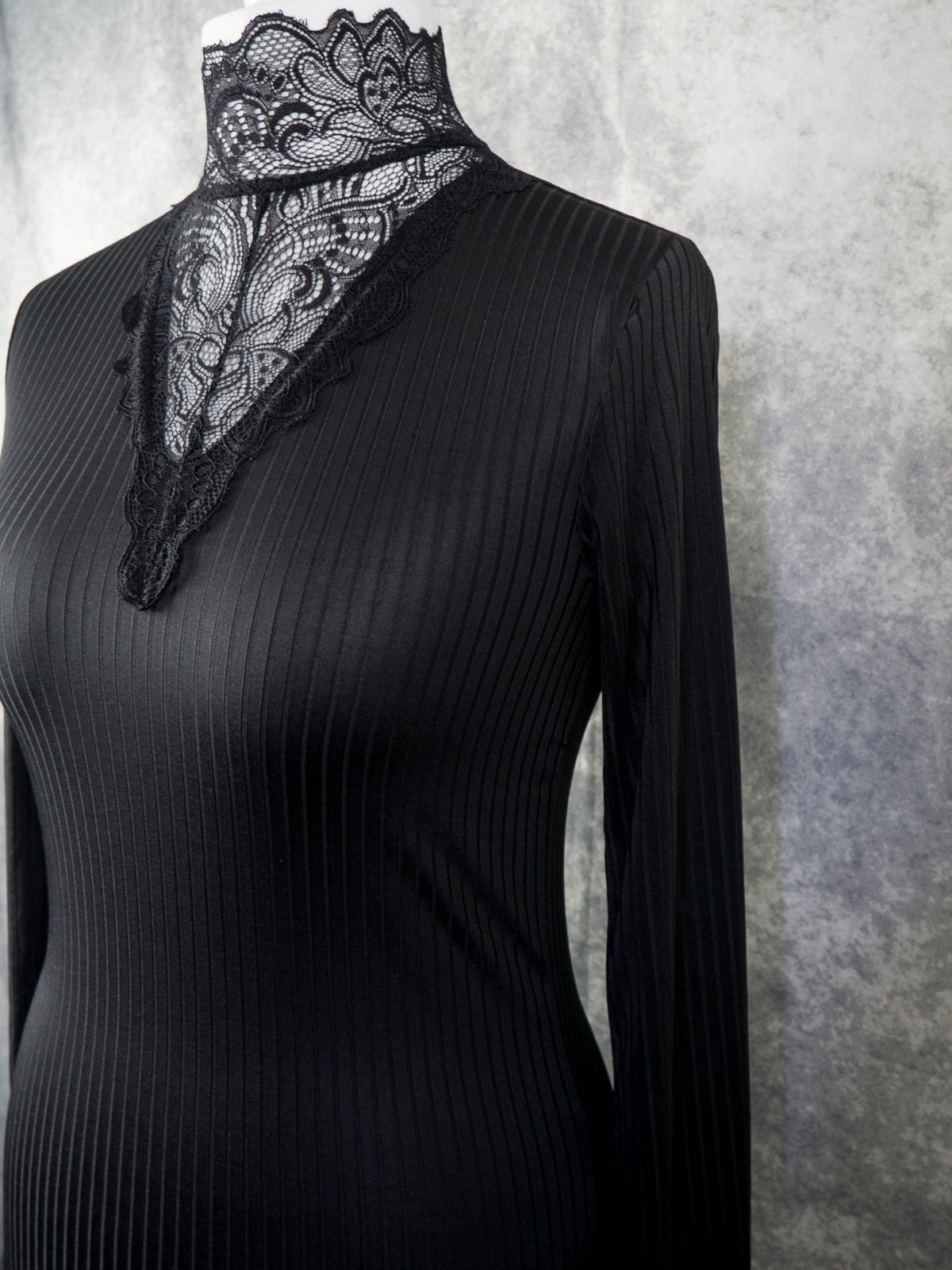A black ribbed long-sleeve top with a high mock neck and a prominent black lace overlay at the neckline, displayed on a mannequin against a textured gray background.