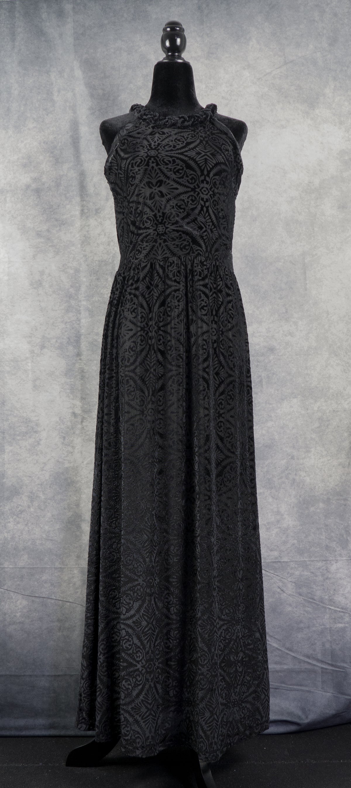 A sleeveless halter-neck maxi dress in black burned velvet (devoré) featuring an intricate, symmetrical damask floral pattern throughout. The floor-length dress is displayed on a black headless mannequin against a textured grey background.