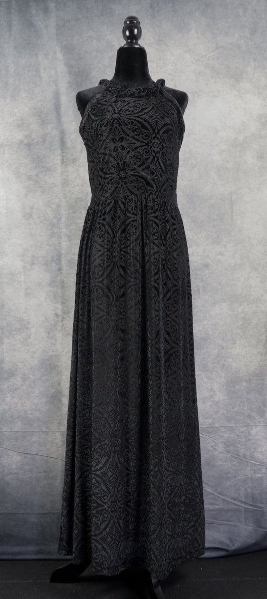A sleeveless halter-neck maxi dress in black burned velvet (devoré) featuring an intricate, symmetrical damask floral pattern throughout. The floor-length dress is displayed on a black headless mannequin against a textured grey background.