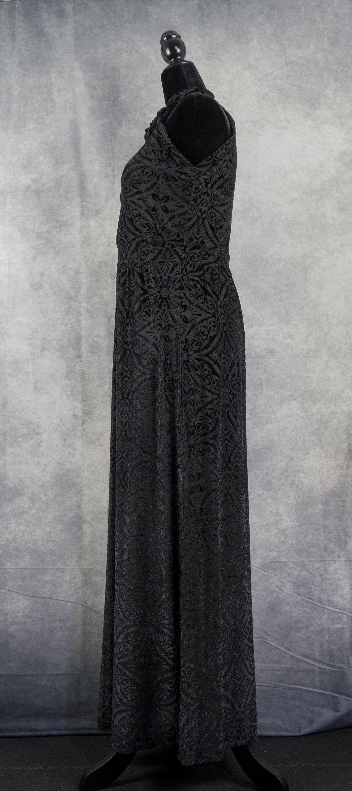 Side profile of a black sleeveless halter-neck maxi dress in devoré velvet with an intricate symmetrical pattern, displayed on a headless mannequin against a textured grey studio backdrop.