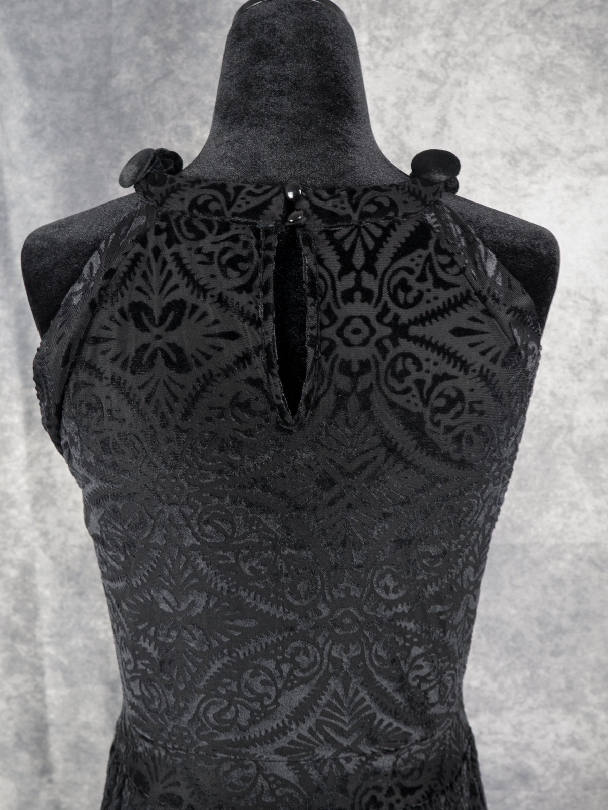Close-up of the upper back of a black devoré velvet halter dress on a mannequin. It highlights a keyhole opening at the neckline, secured with two small black buttons, showcasing the intricate symmetrical burnout floral pattern.