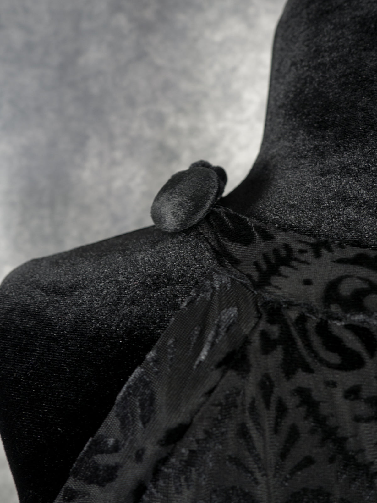 A detailed macro shot focusing on a round, fabric-covered button on the shoulder strap of the black devoré velvet halter dress, displayed on a black velvet mannequin.