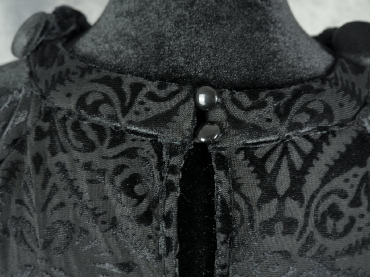 An extreme close-up of the back neckline of the black devoré velvet dress, showing the details of two small, glossy black buttons fastening the top of the keyhole closure.
