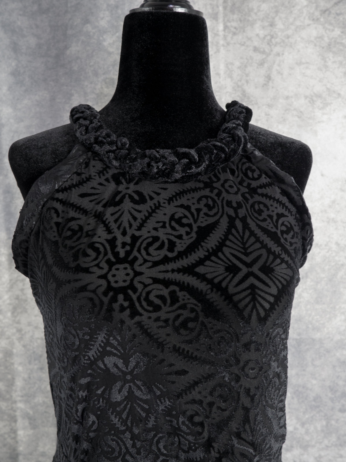 A close-up shot of the front bodice and neckline of the black devoré velvet halter dress. It highlights the ruched, braided-style halter neckline trim and the intricate, symmetrical damask-inspired burnout pattern on the chest.