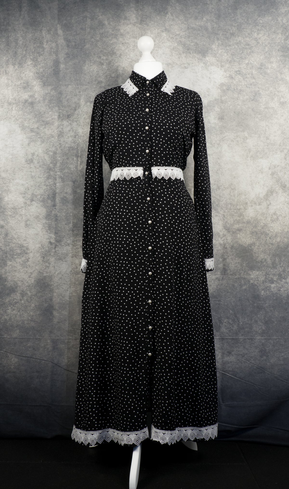 A long-sleeved, full-length black maxi dress with a white polka dot pattern displayed on a white mannequin. The dress features a pointed collar, white pearl-like buttons running down the entire front center, and decorative white crochet lace trim detailing the collar, waistline, cuffs, and bottom hem. The backdrop is a textured grey studio fabric.