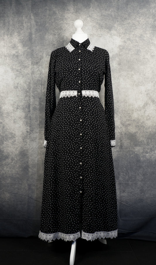 A long-sleeved, full-length black maxi dress with a white polka dot pattern displayed on a white mannequin. The dress features a pointed collar, white pearl-like buttons running down the entire front center, and decorative white crochet lace trim detailing the collar, waistline, cuffs, and bottom hem. The backdrop is a textured grey studio fabric.