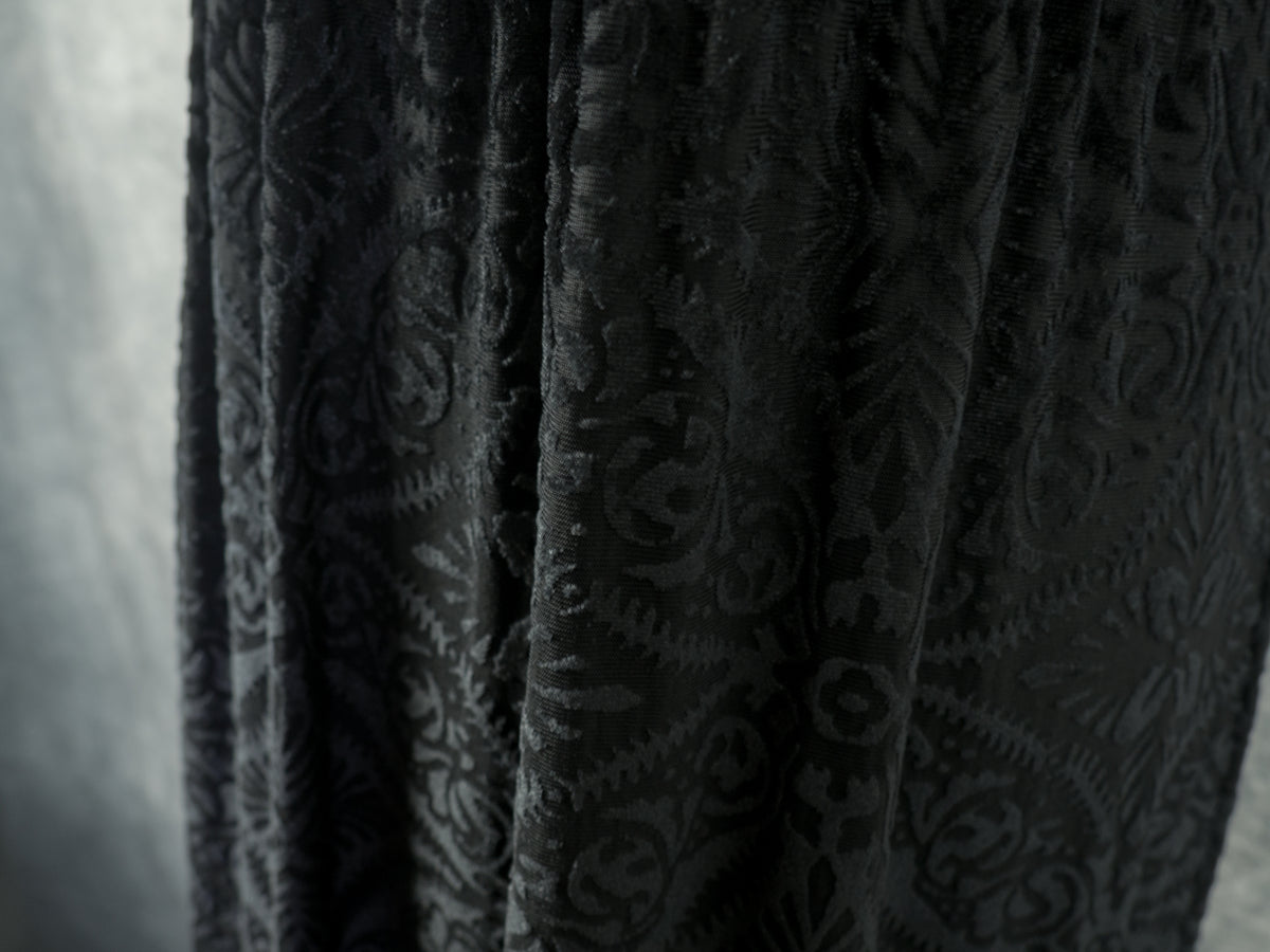 A close-up shot of the dress's skirt fabric, showing the natural folds and drapes of the black devoré velvet that capture the light and emphasize the texture of the damask pattern.