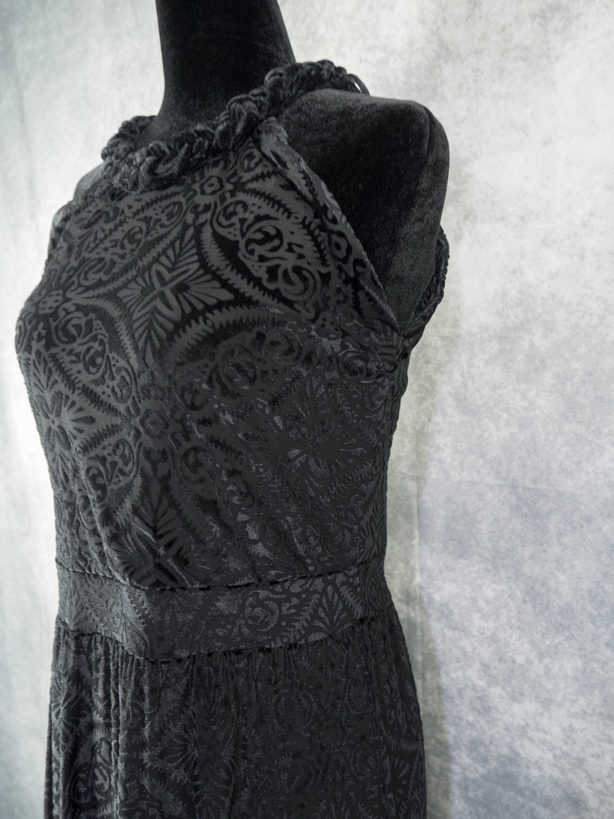 A three-quarter side view of the upper bodice of the black devoré velvet halter dress on a mannequin. It showcases the fitted waist seam, the braided neckline trim, and the intricate damask burnout pattern extending across the torso.