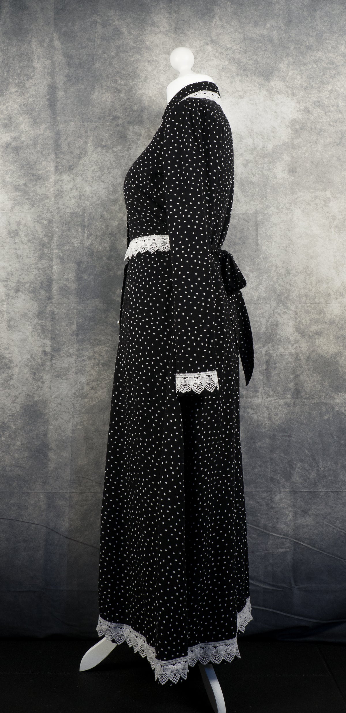 Side view of a long-sleeved black maxi dress with a white polka dot pattern shown on a mannequin. A fabric belt is tied into a bow at the back of the waist. The white crochet lace trim is visible on the collar, waistline, cuffs, and scalloped bottom hem against a textured grey background.