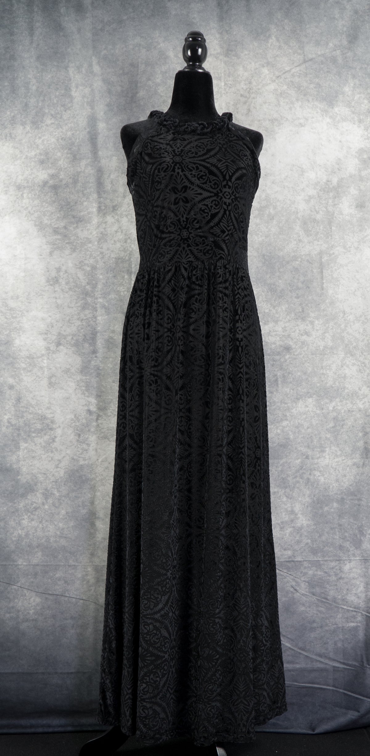 "A full-length front view of the black sleeveless halter-neck maxi dress on a black headless mannequin. The floor-length dress displays a continuous, intricate damask floral burnout pattern across the entire devoré velvet fabric, set against a mottled grey background.