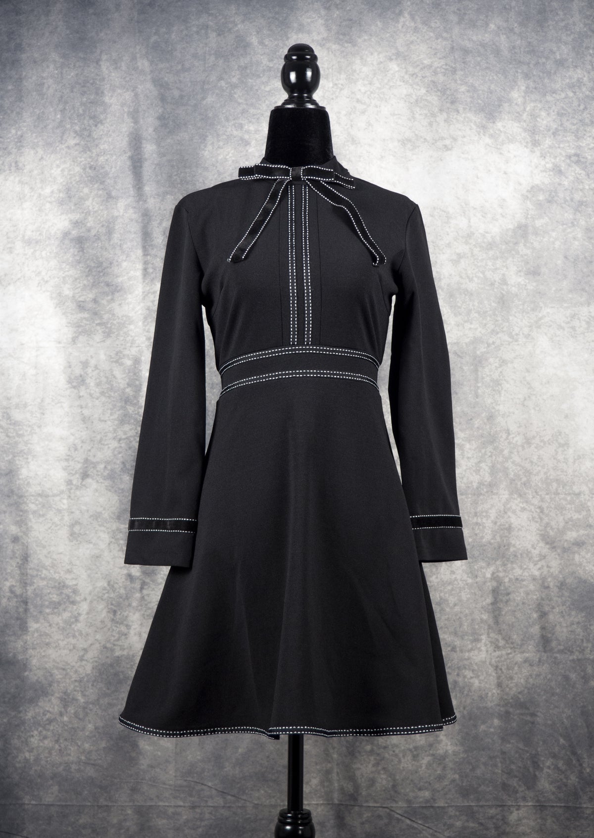 Full front view of a black long-sleeve gothabilly dress on a mannequin, featuring a bow tie neckline, white contrast topstitch details, and a vintage fit-and-flare A-line silhouette.
