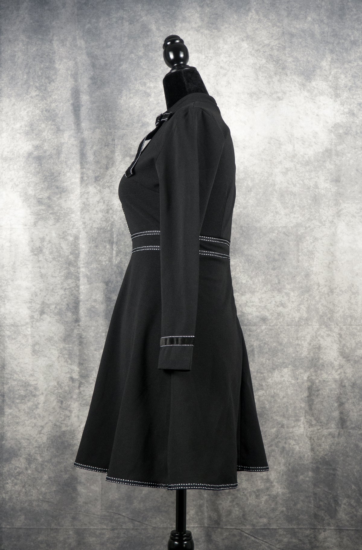 Side view of a black long-sleeve gothabilly dress on a mannequin, featuring a bow tie neckline, white contrast topstitch details, and a vintage fit-and-flare A-line silhouette.
