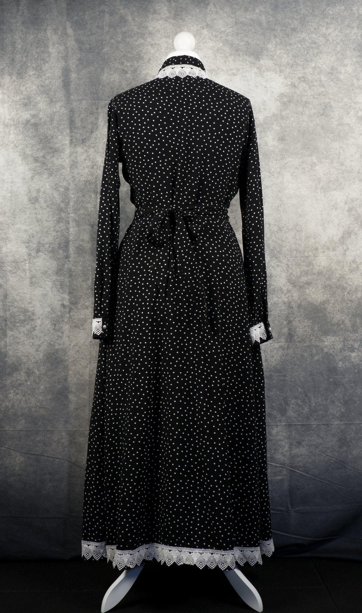 Back view of a long-sleeved black maxi dress with a white polka dot pattern on a mannequin. The dress features a fabric sash tied into a neat bow at the center of the waist. White crochet lace trim accentuates the back of the collar, the sleeve cuffs, and the scalloped bottom hem against a textured grey background.