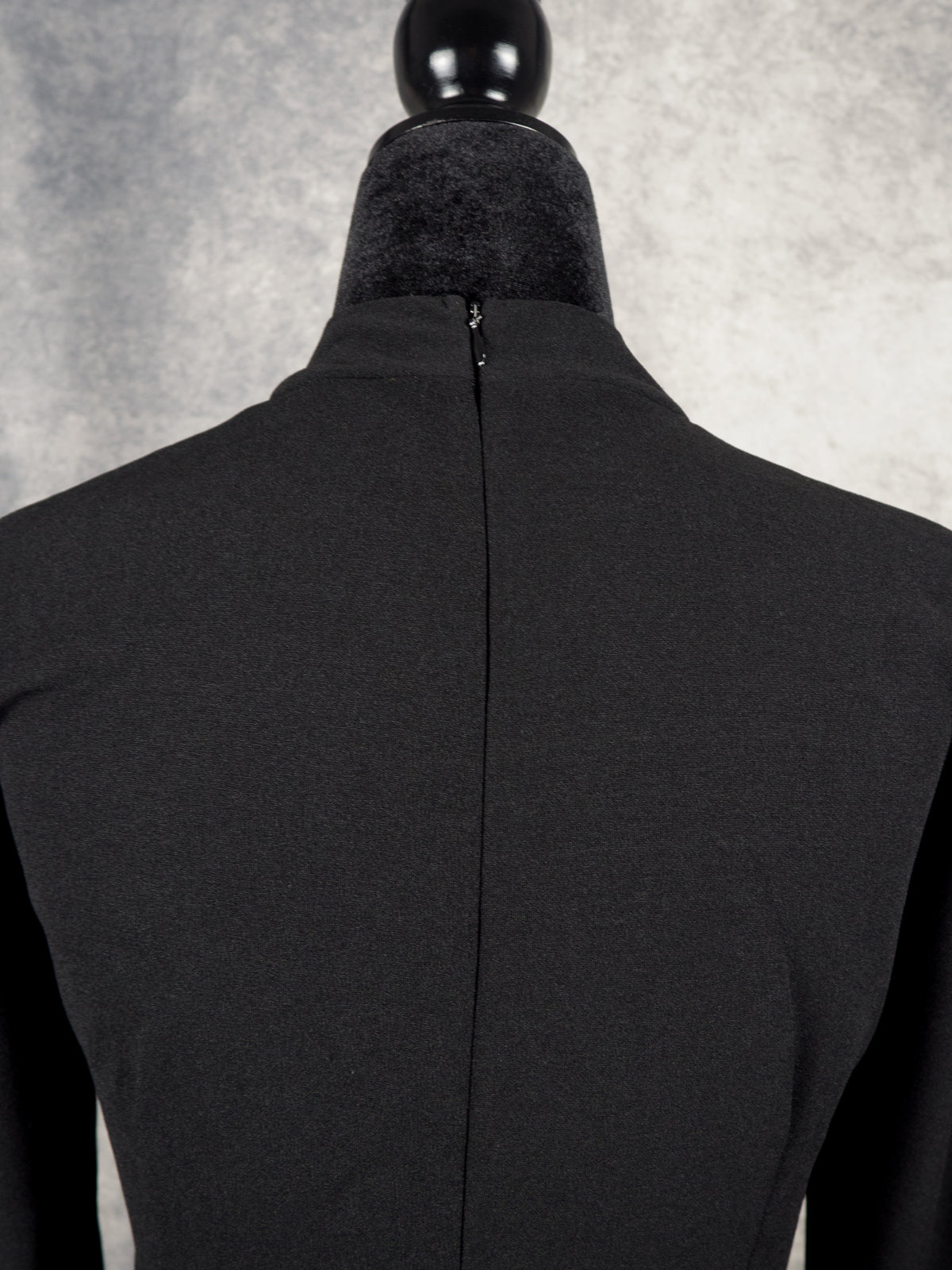 Back view close-up of a black high-neck gothabilly dress on a mannequin, highlighting the hidden vertical zipper closure, textured black fabric, and alternative retro styling.

