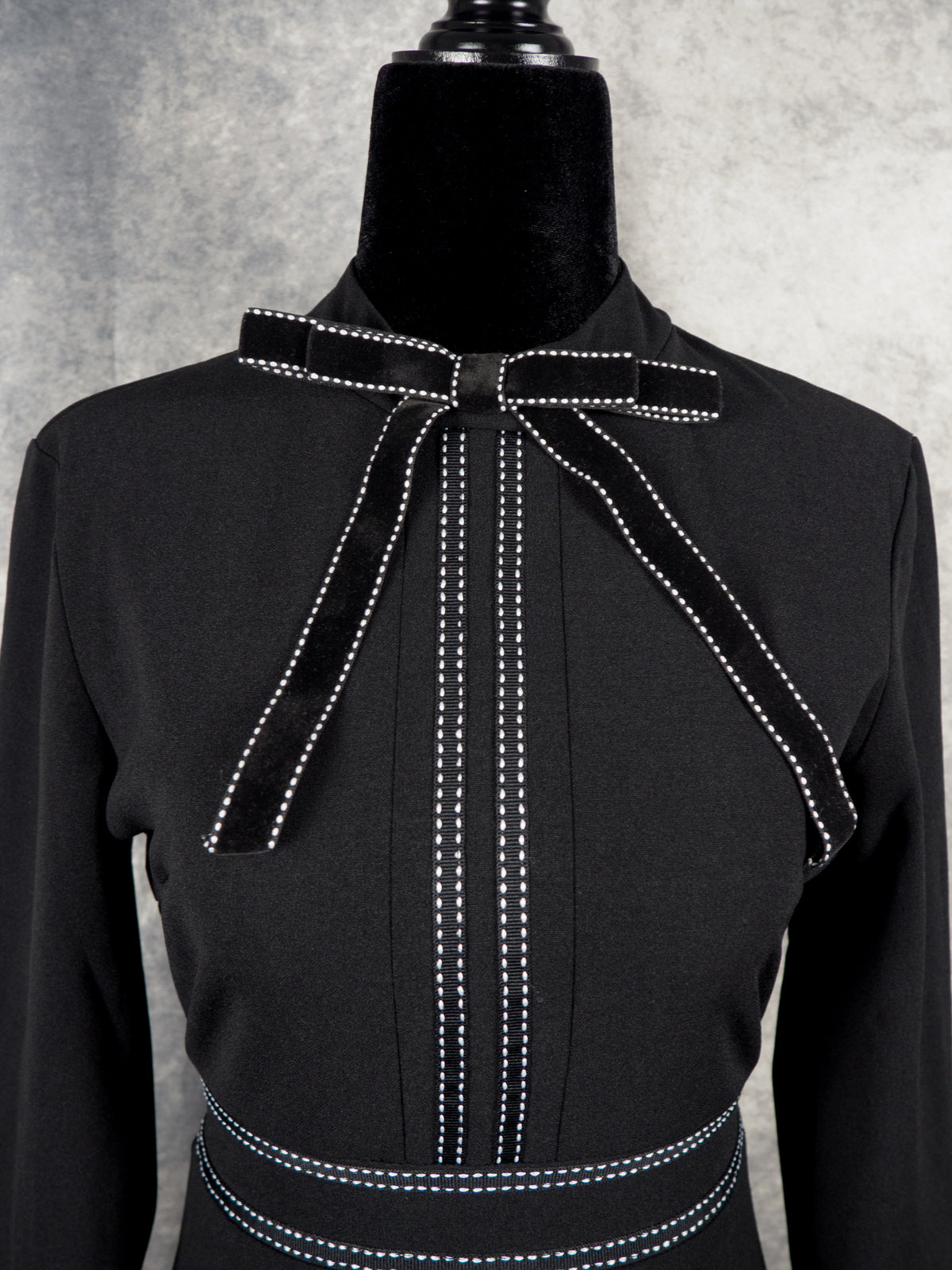 Close-up of the neckline on a black alternative gothabilly dress, highlighting a black velvet bow tie collar and vertical white contrast topstitch details down the bodice.
