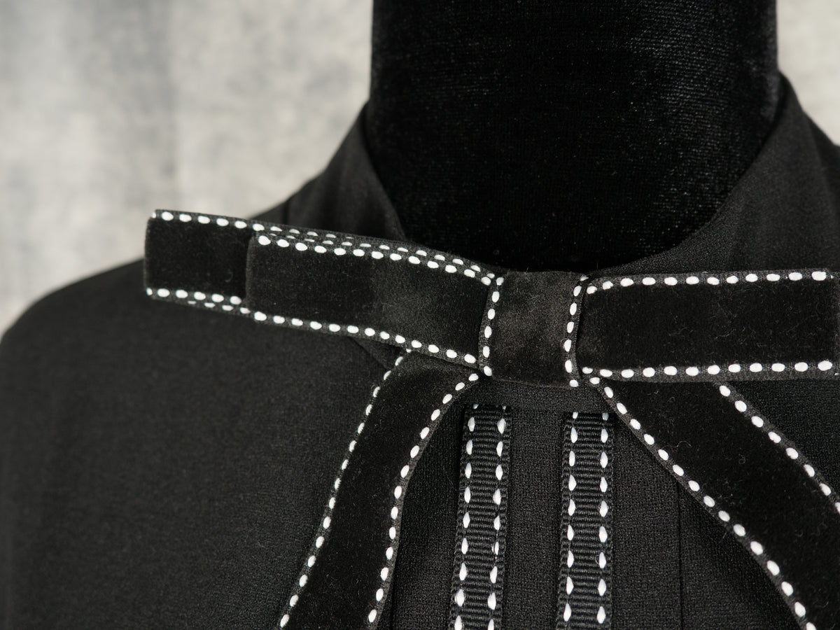 Extreme close-up of a black velvet bow on an alternative gothabilly dress collar, showcasing intricate white contrast pinstitch details and textured fabric on a mannequin.
