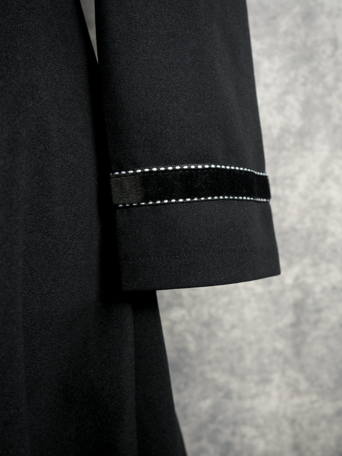 Close-up of a black dress sleeve with white contrast stitching, set against a textured gray background.