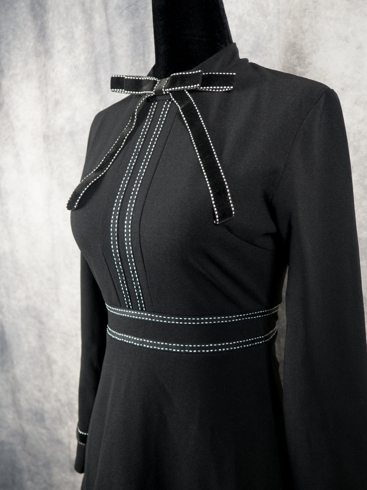 Three-quarter front view of a black alternative gothabilly dress on a mannequin, showcasing the bow tie collar, structured bodice, and white contrast topstitch panels.
