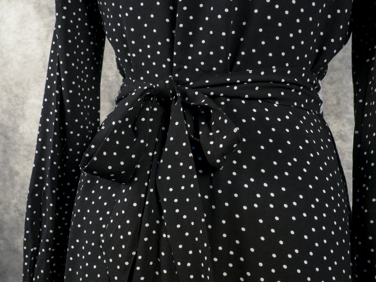 Close-up of the waist area of a black garment with a white polka dot pattern. A matching fabric belt is tied into a detailed bow at the waistline. The long sleeves of the garment drape down on either side against a blurred, textured grey background.