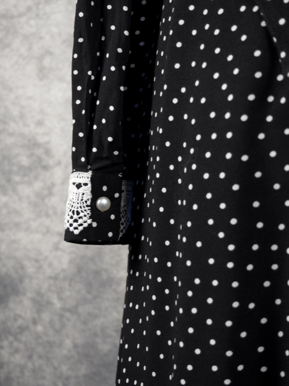 Close-up of the sleeve cuff of a black garment with a white polka dot pattern. The cuff is fastened with a round white pearl-like button and detailed with a patch of intricate white crochet lace. The background is a textured grey wall.