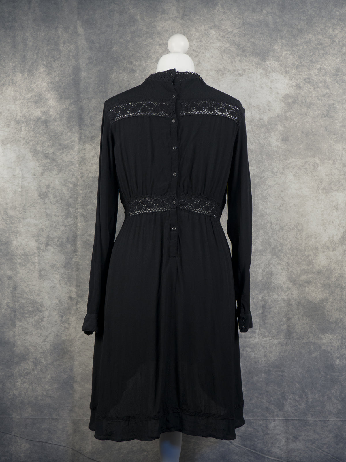A full back view of a long-sleeved black mini dress on a white mannequin. It features a high lace-trimmed collar and a vertical row of small buttons running down the center back. Semi-sheer geometric lace panels are horizontally inlaid across the upper shoulders and the defined empire waistline, contrasting with a textured grey studio background.