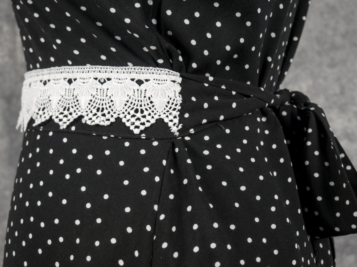Close-up of the waistline of a black garment with a white polka dot pattern. A matching fabric sash wraps around the waist, featuring a prominent section of decorative white crochet lace trim at the front, and is tied into a knot or bow at the side. The background is a textured grey studio surface.