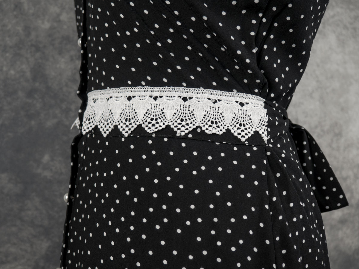 Close-up of the waist area of a black button-front dress with a white polka dot pattern. A wide fabric belt featuring an intricate white crochet lace trim with scalloped edges is wrapped around the waistline. A portion of a fabric bow is visible tied at the side against a textured grey background.