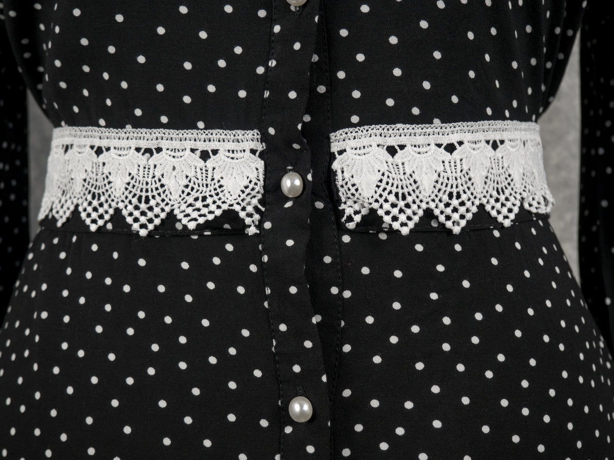 Front close-up of the waist area of a black button-down dress featuring a white polka dot pattern. The dress is fastened down the center with round white pearl buttons, which divide a matching fabric belt adorned with broad, intricate white crochet lace trim.