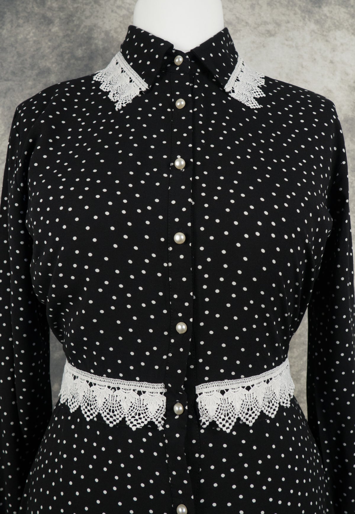 Close-up of the upper bodice of a black long-sleeved dress with a white polka dot pattern. The dress features a pointed collar accentuated with white crochet lace trim at the tips and a series of round, white pearl buttons running down the center placket. A matching fabric belt with matching white lace trim defines the waistline.