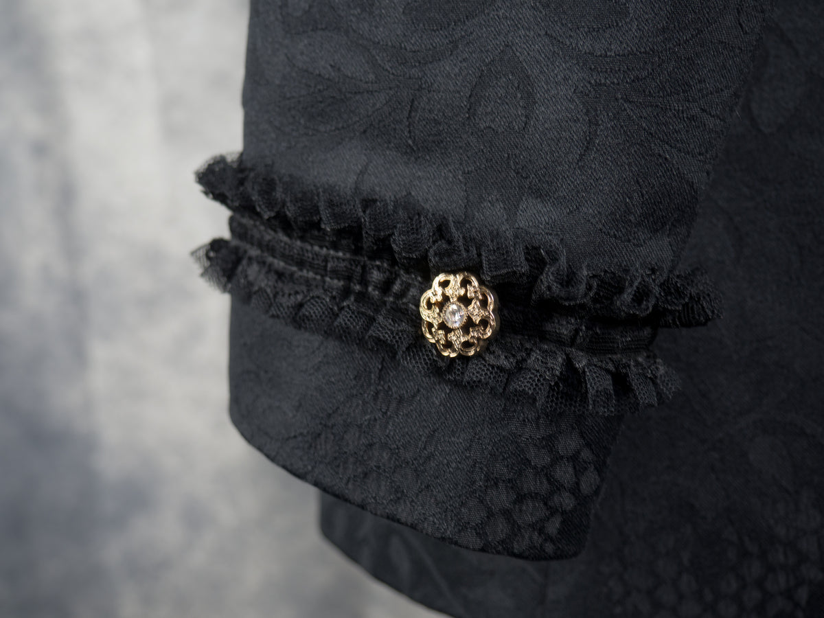 A close-up of a black textured fabric garment with ruffled trim and a decorative gold-toned brooch featuring a clear central stone.