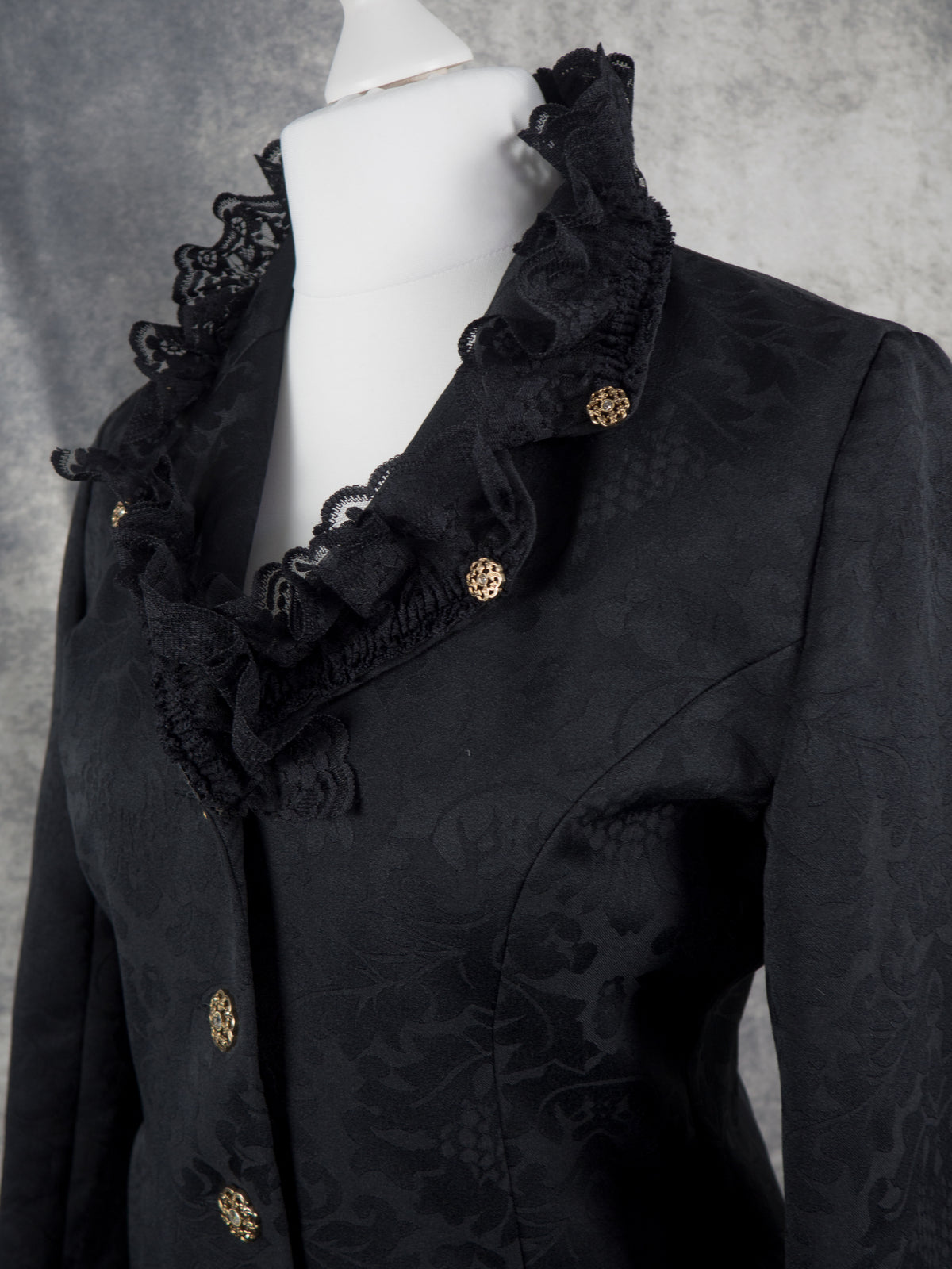 A black jacquard blazer with ruffled lace trim and decorative gold buttons, displayed on a white mannequin torso against a textured gray background.